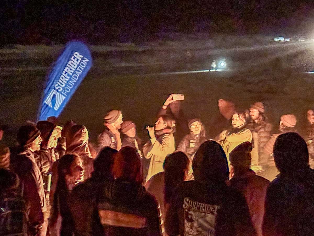Small crowd gathered around a bonfire on the beach with the Surfrider flag visible and illuminated by the fire on Asilomar Beach Monterey, Ca.. Photo: Harry Parker Photography