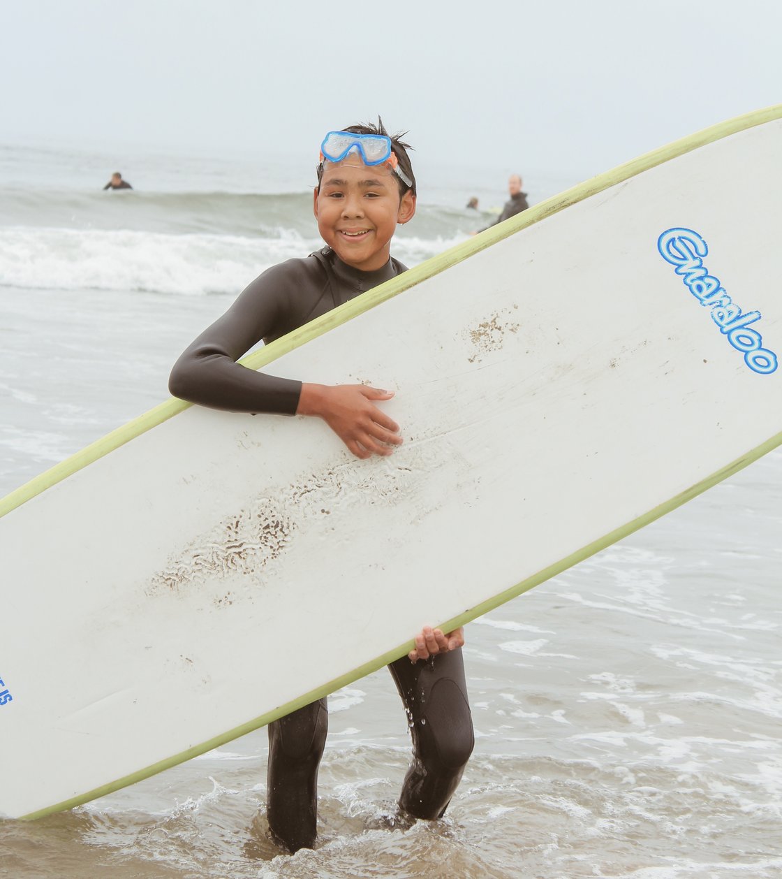 a child smiling and holding a surf board at a youth surf camp at Bastendorff Beach