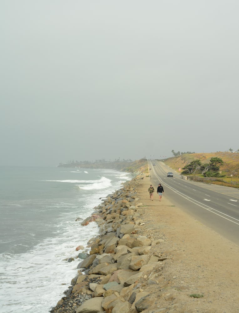 Image shows rip rap (large rocks) piled up between a vehicle road and the ocean. The tide is high and completely covering the beach leaving no sand or beach area to access.