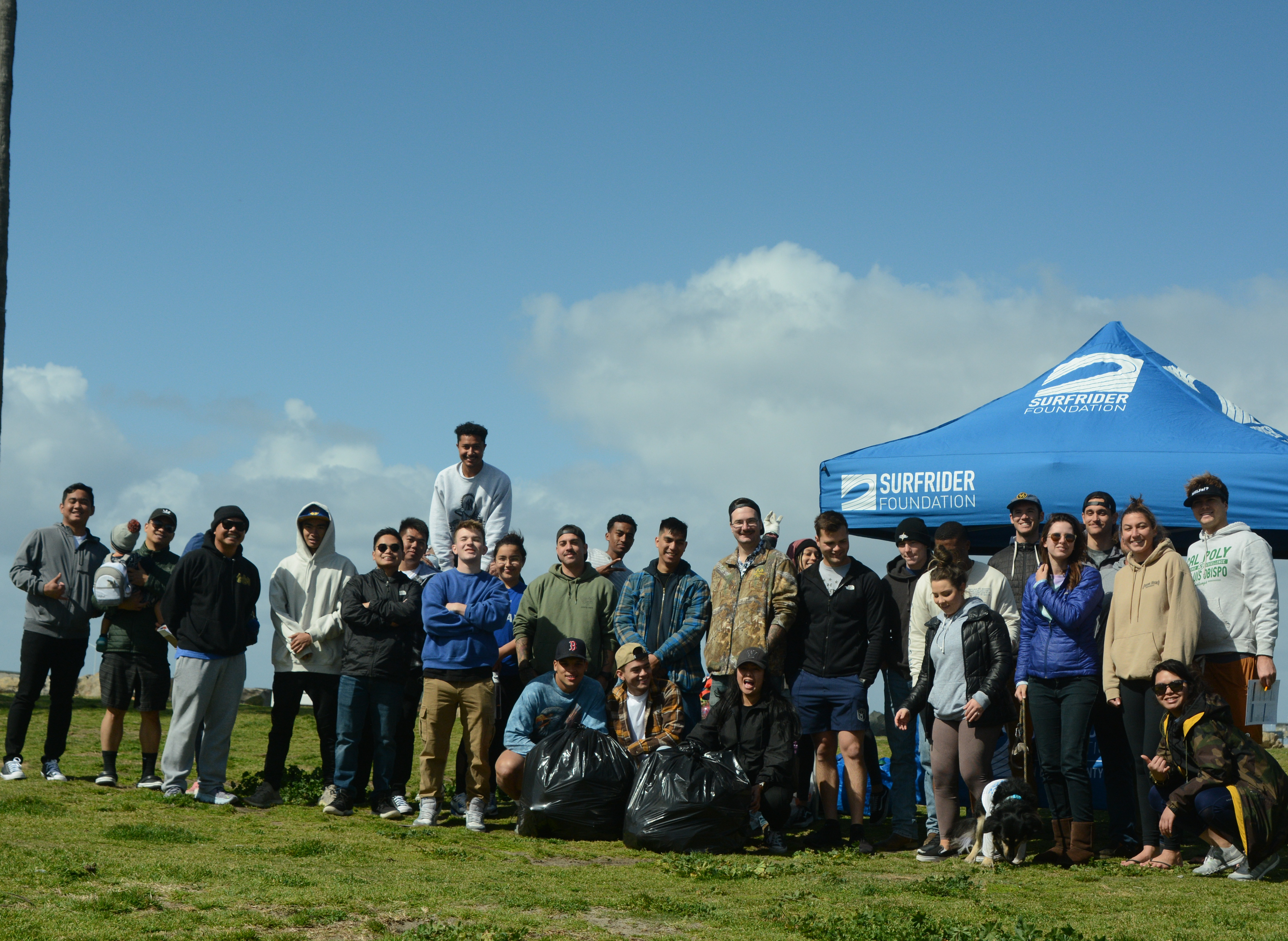 A large group of people standing on grass in front of a beach, posing with three large bags of trash that were collected off the beach.
