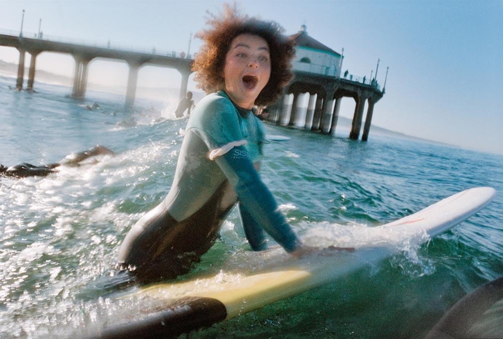 Photo of a woman on a surfboard smiling in the ocean taken by Gabriella Angotti-Jones