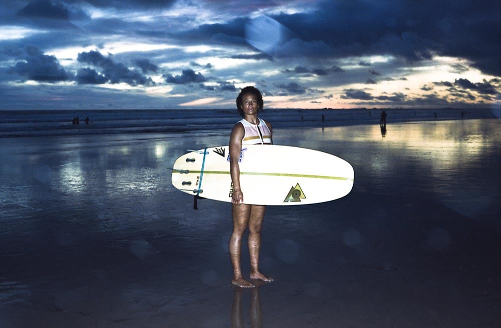 Photo of a woman standing on a beach holding a surfboard at sunset taken by Gabriella Angotti-Jones