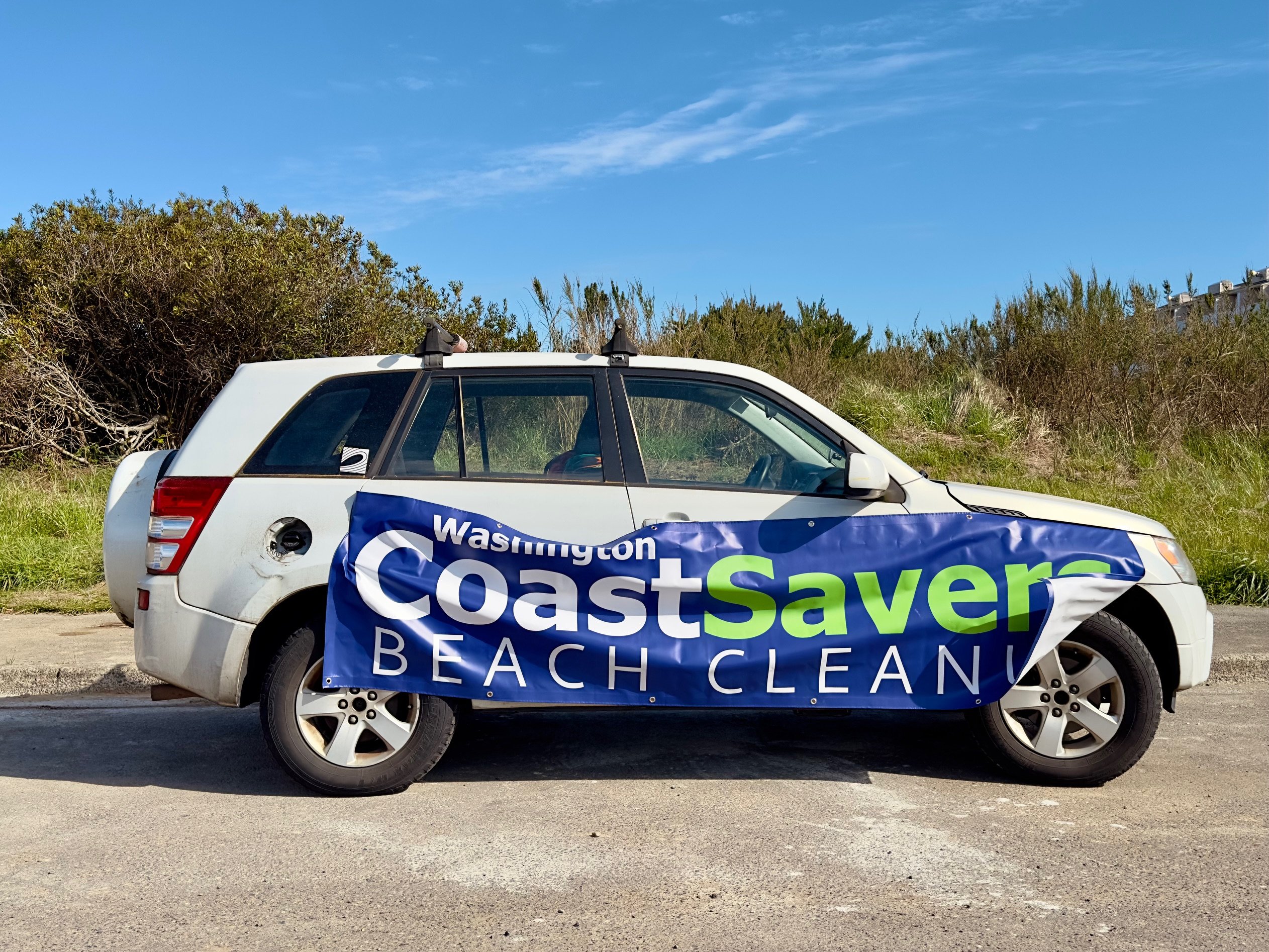 The Surfrider Suzuki holding up a large Coastsavers Beach Cleanup banner in front of dune scrub under blue skies