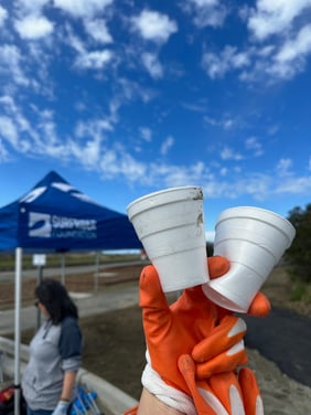 A gloved hand holds up two used styrofoam cups in front of a Surfrider beach cleanup tent under blue skies 