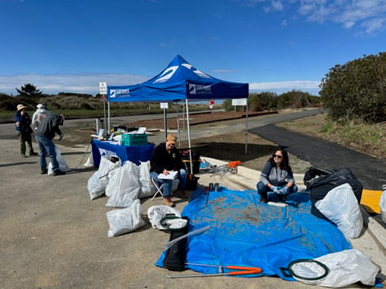 Grays Harbor chapter volunteers sit in front of a Surfrider canopy, ready to sort trash under blue skies