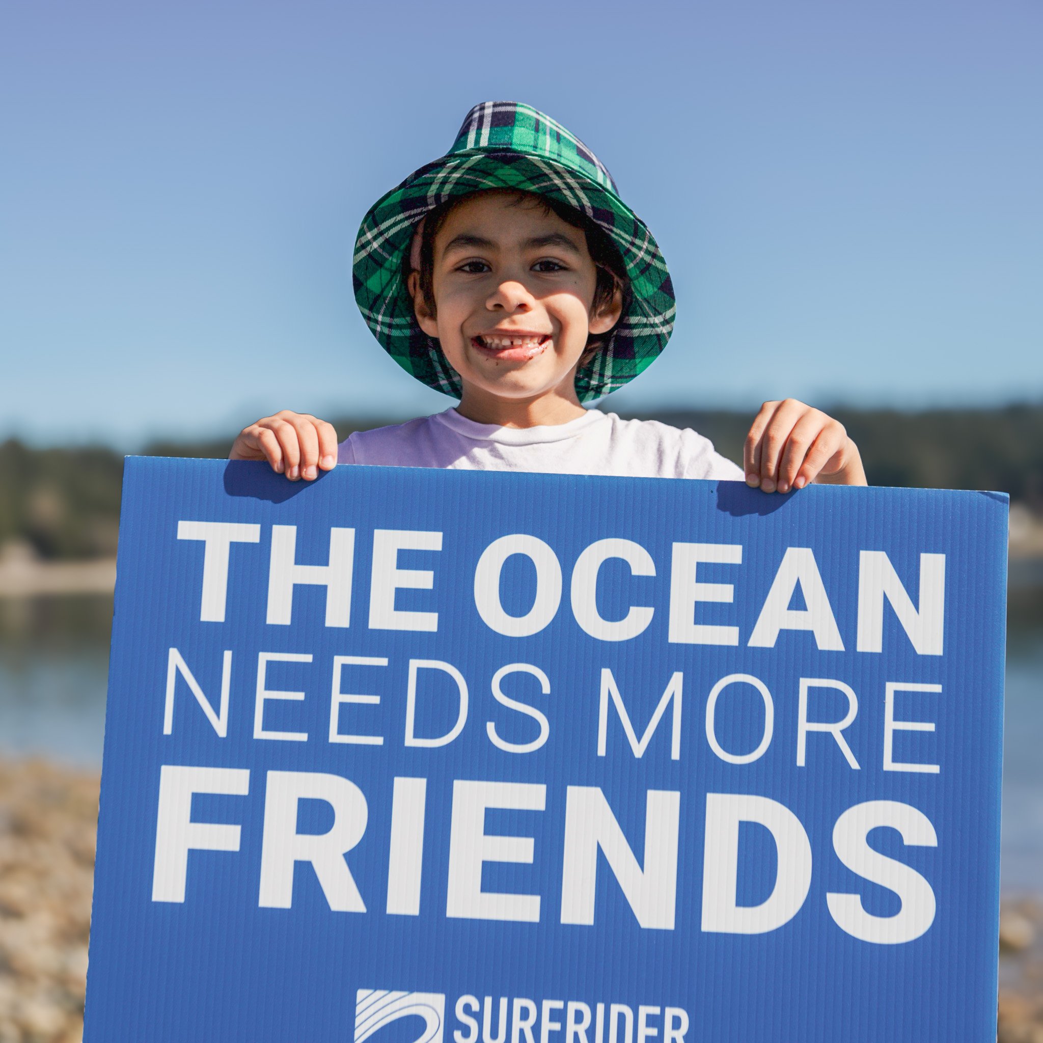 A young boy wearing a plaid green hat, smiling big, and holding a sign that says The Ocean Needs More Friends