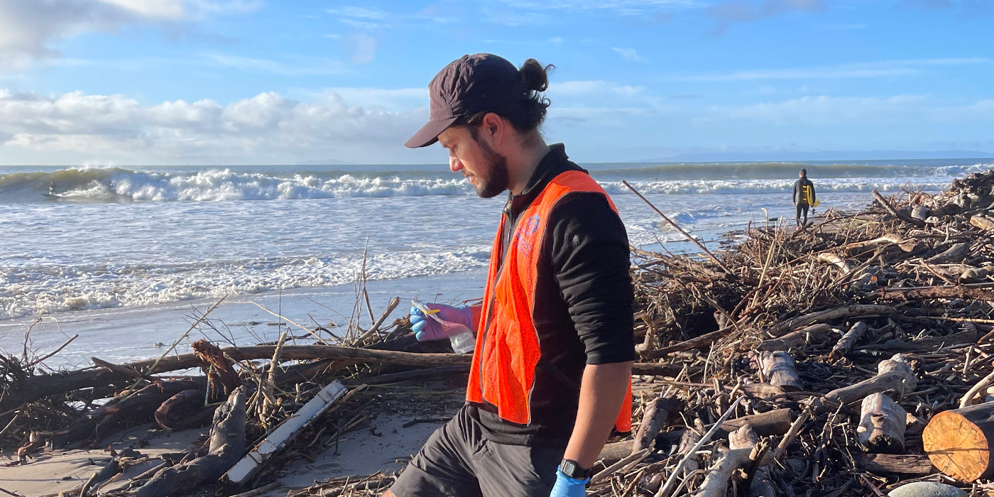 A Blue Water Task Force Volunteer collecting a water sample as surfers enjoy waves in the background, highlighting the importance of water quality testing. The water sample was tested for enterococcus bacteria. 