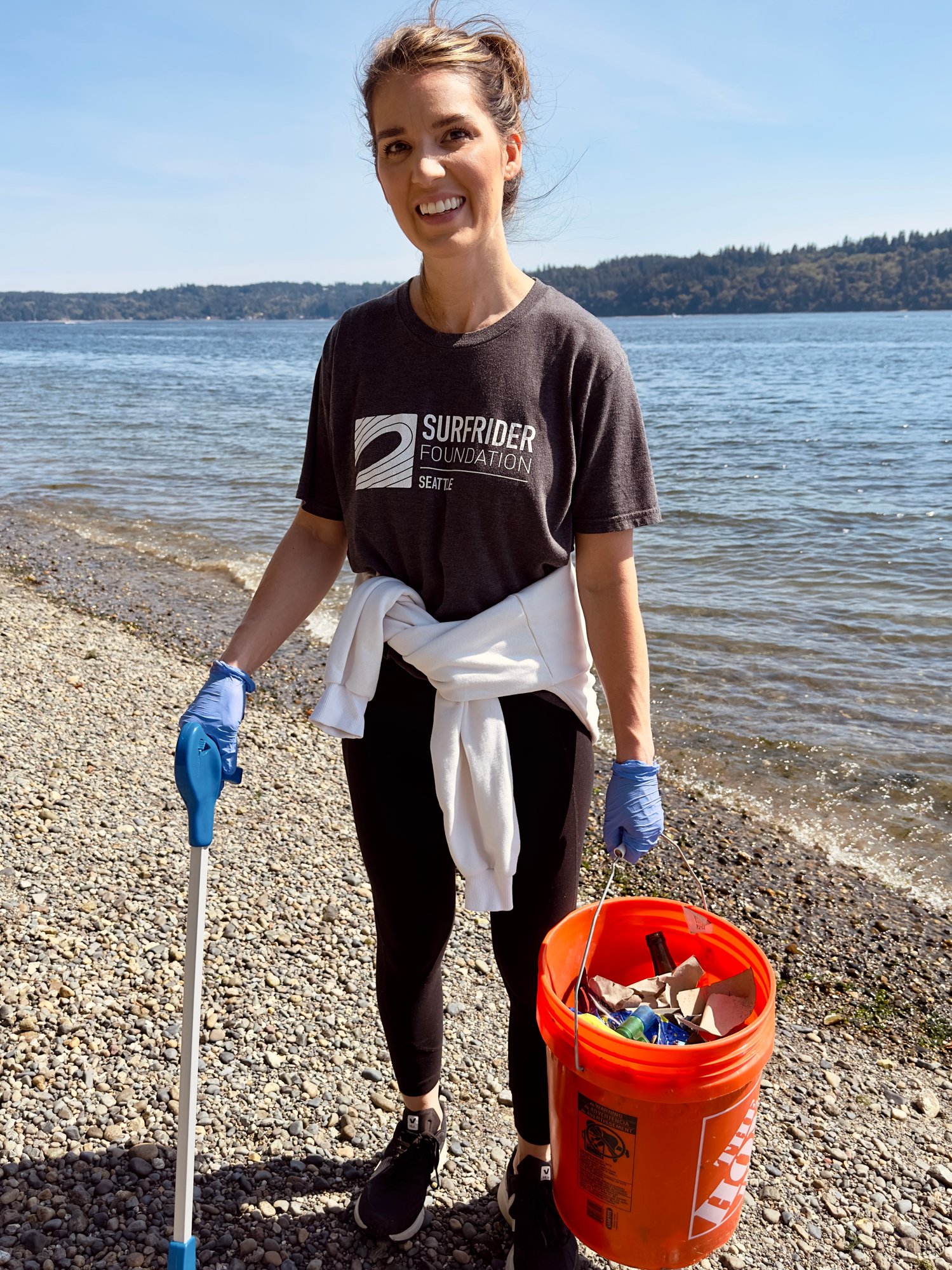 A female volunteer wearing a Surfrider t-shirt smiling and posing with her bucket of trash during a beach cleanup