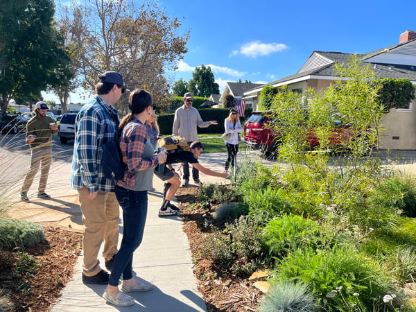 A group of people stand in front of a sunny front yard and talk about plants