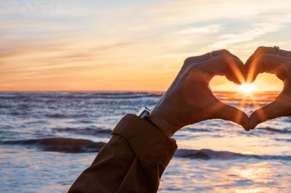 A person making the shape of a heart with their hands and capturing a sun setting over the ocean