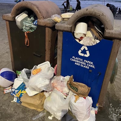 Trash cans overflowing at the beach near Oceanside pier, with piles of trash around the cans.