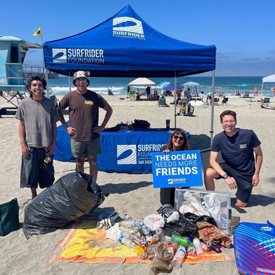 Volunteers gather in front of the Surfrider tent at the Oceanside beach cleanup. Plastic bags, bottles and other debris from the cleanup are laid out on a towel in front of them.