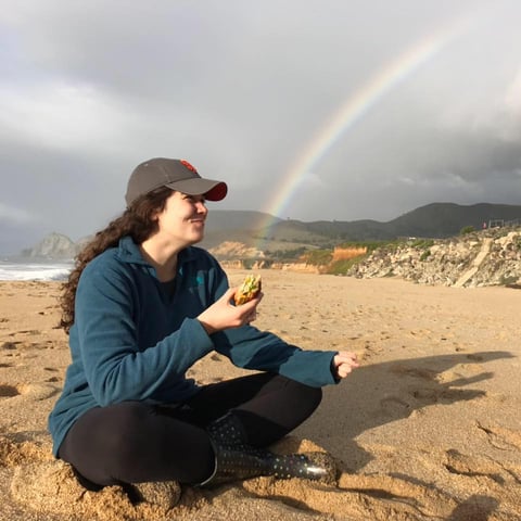 Sarah smiling and eating a sandwich on the beach with a rainbow in the background</p>
<p>