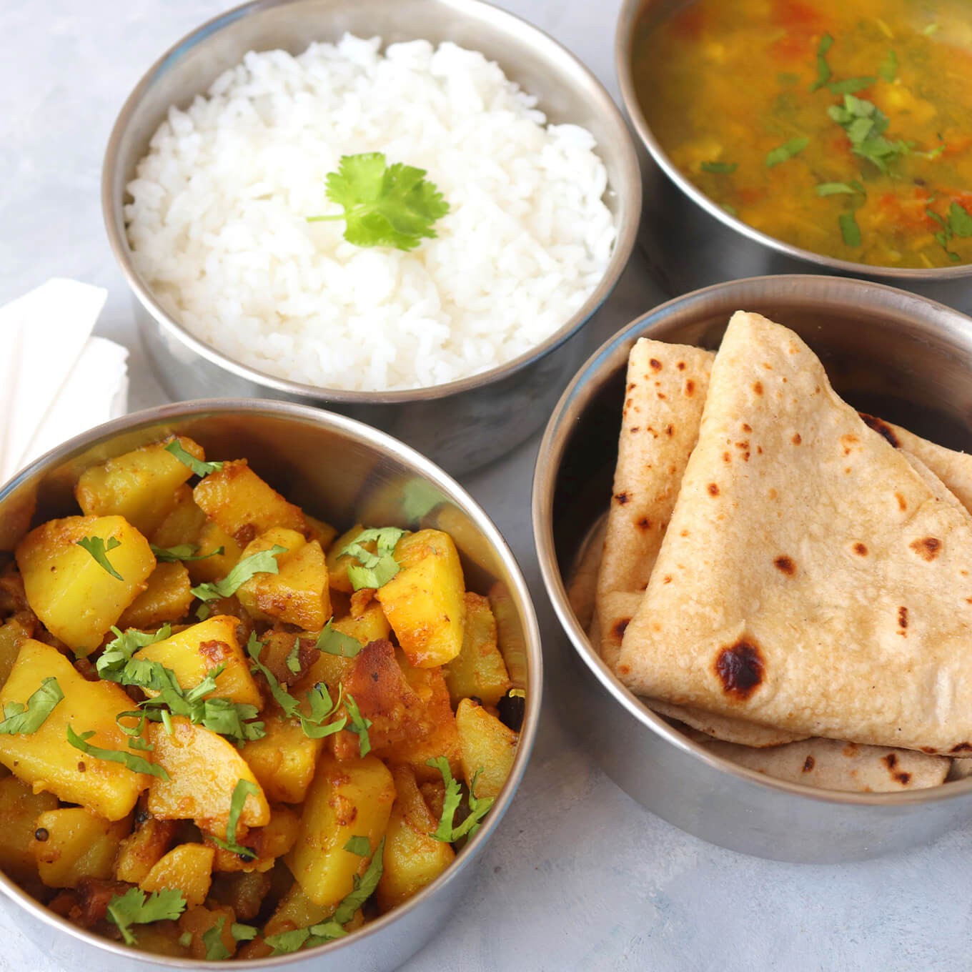 An overhead shot of 4 metal round food containers, tuffins, holding brigth potatoes covered in an orange sauce, another container holds rice with a geen leaf in the middle, another holds flat bread triangles, and the last container holds a red and green soup or curry.