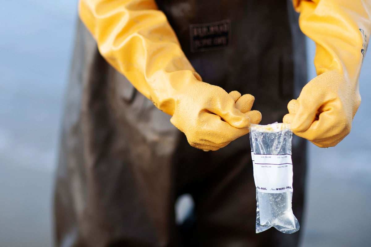 A figure in protective gear holds a sample of collected seawater to be sent to testing.