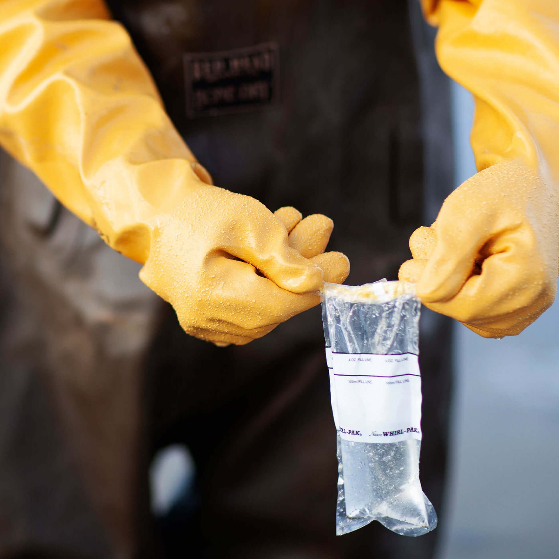 Hands with yellow gloves holding a water test sample in a plastic bag