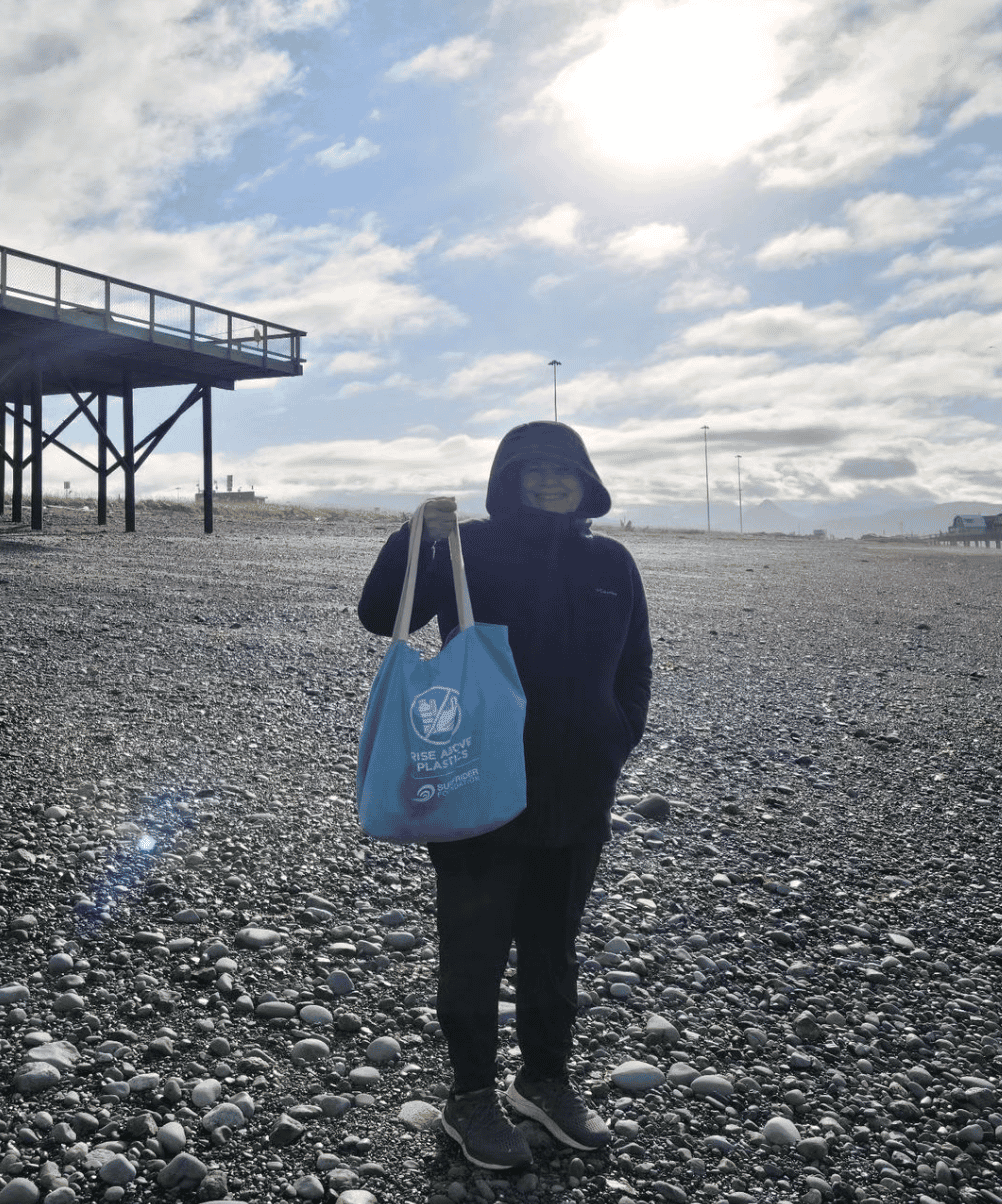 A volunteer holds up a bag full of trash on a pebble beach in front of a pier on a cold, sunny day