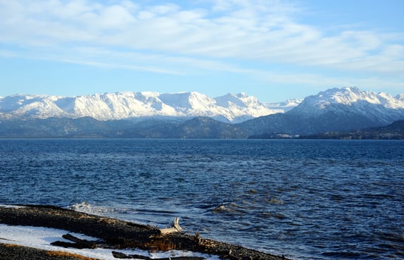 A rugged shoreline with snow capped mountains off the horizon on a blue sky day