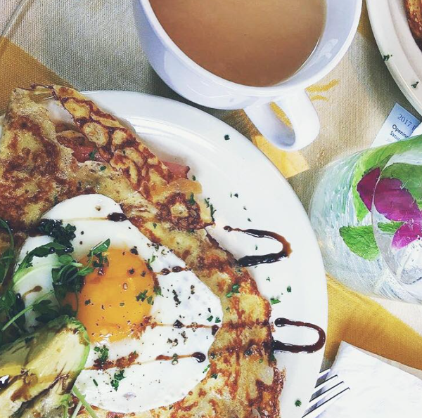 A crepe with a fried egg, avocado, and drizzle posed next to a cup of coffee and water with mint and a purple flower