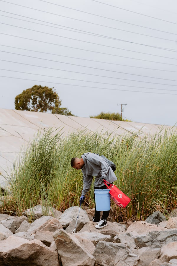 Man wearing gray jacket holding white bucket and red bag is bent over in river bed picking up trash