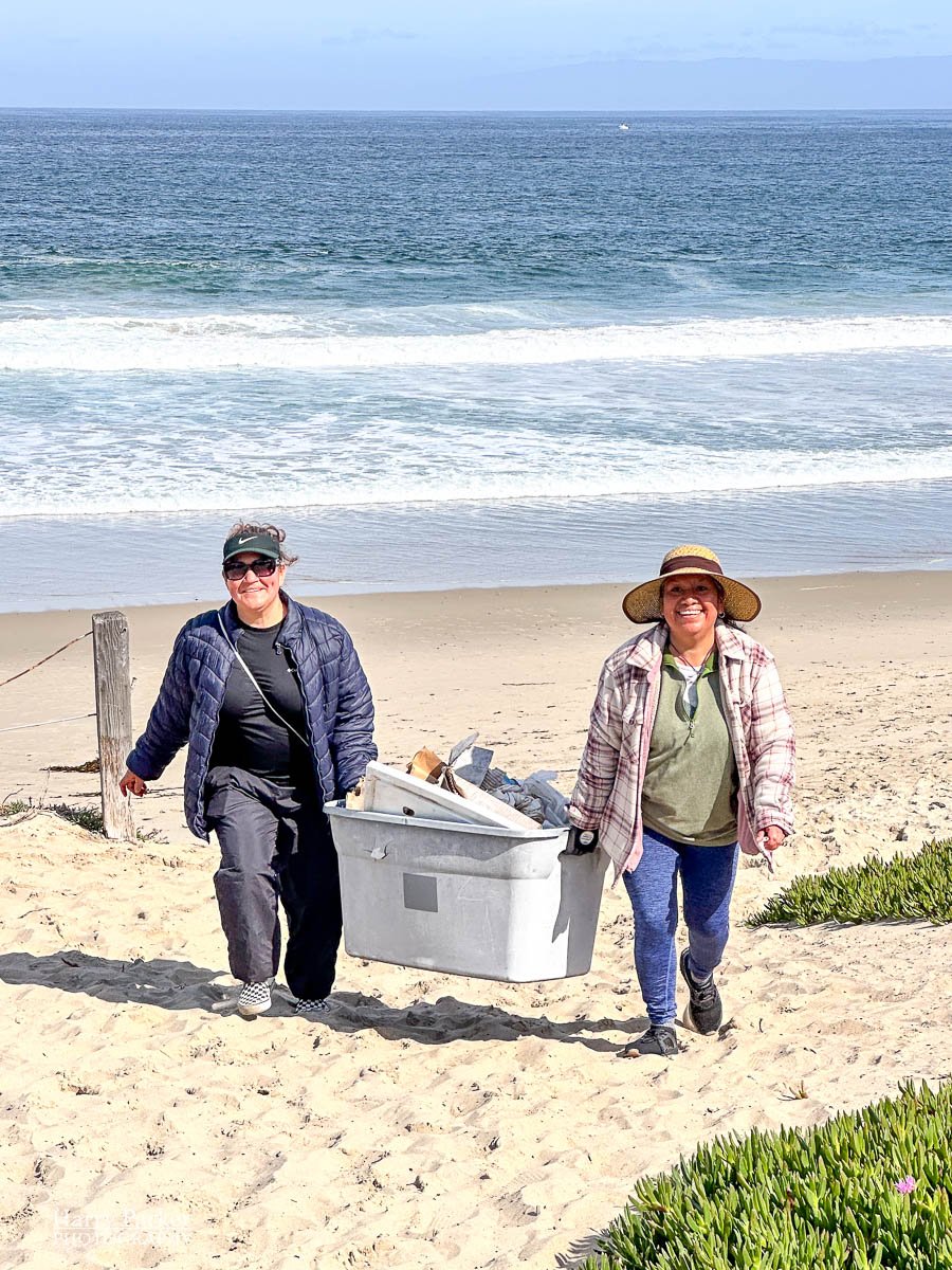 Two women carry a bin full of garbage they plucked form the beach, the waters edge in the background.
