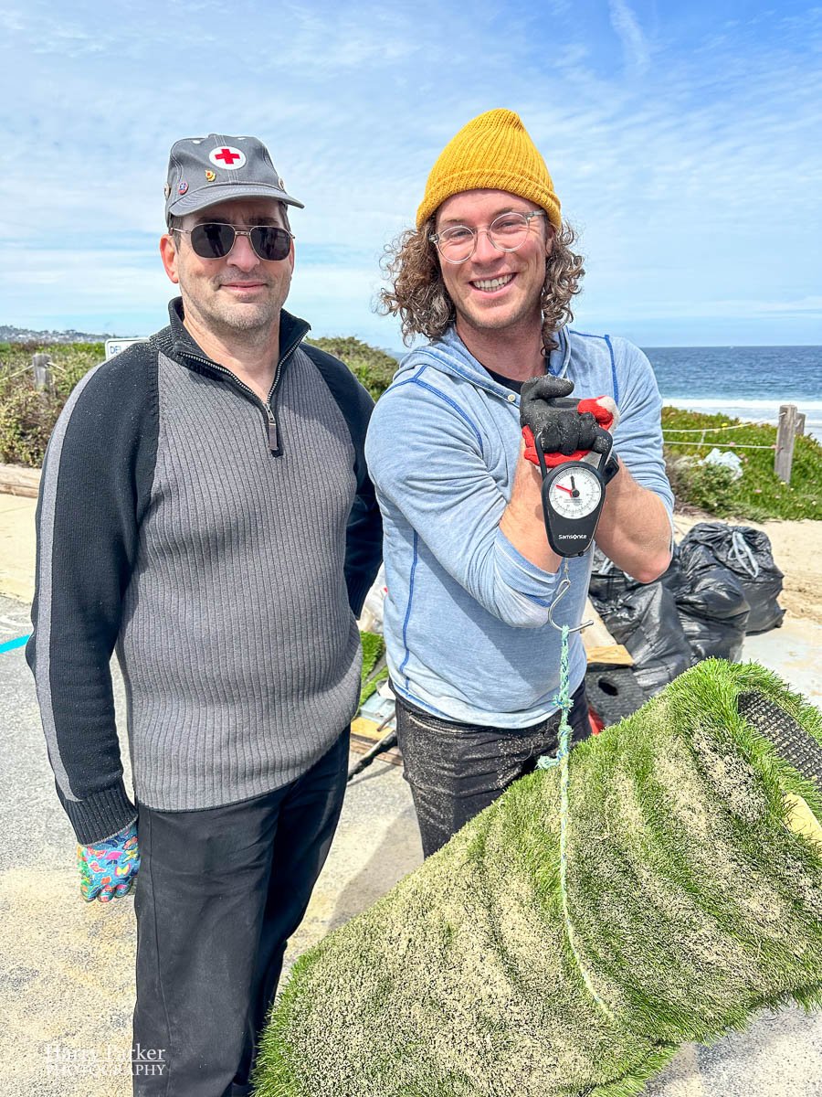 two men weigh a piece of garbage from the beach with smiles.