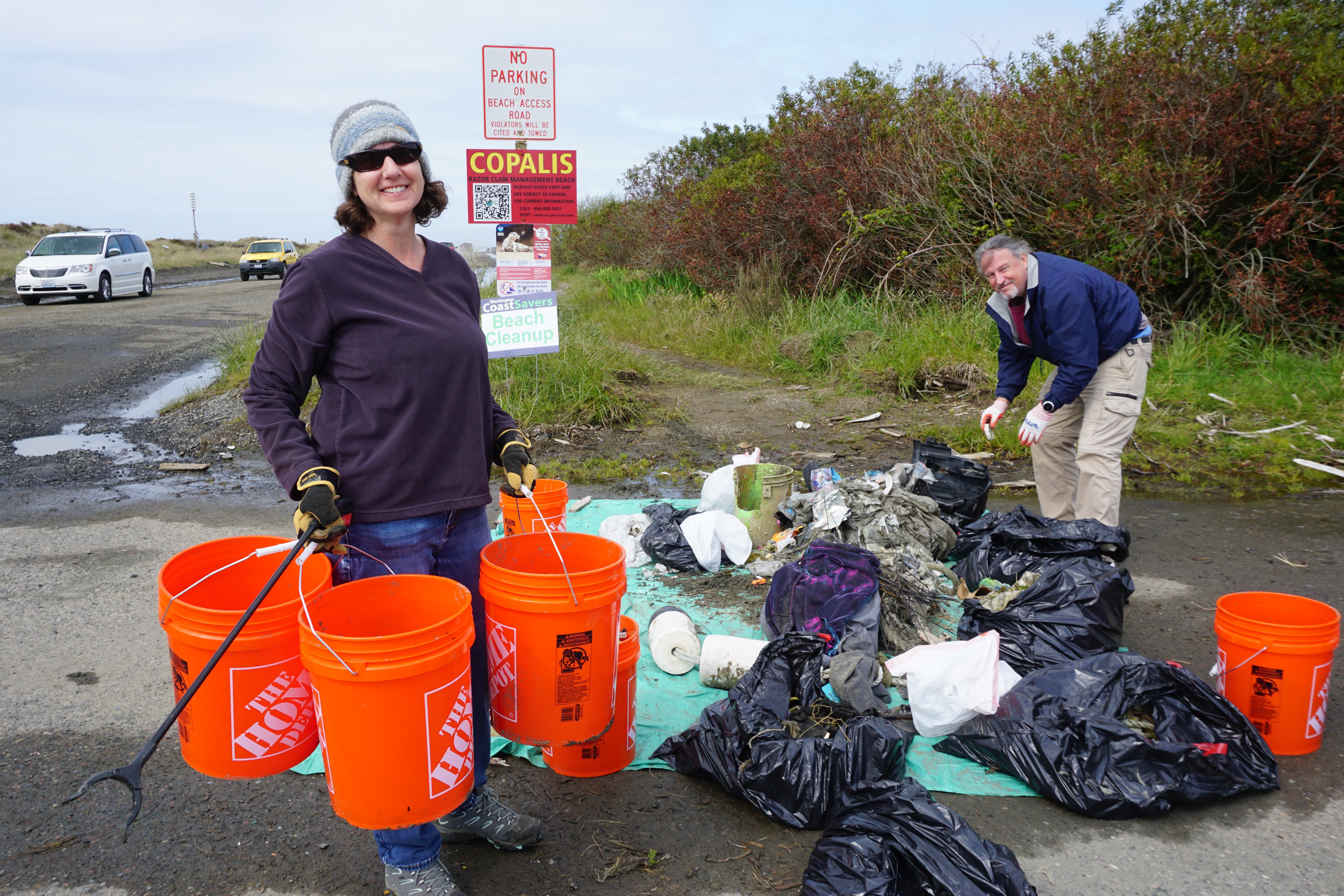A man sorts trash into bags while a woman carries three orange buckets full of debris off the beach