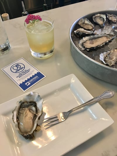 A dozen fresh oysters on the halfshell sitting next to a bright colored cocktail and an OFR sticker