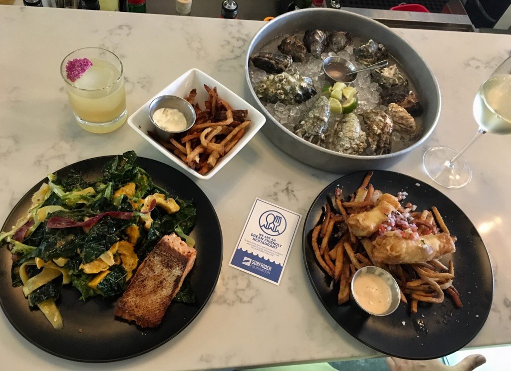 A smorgasbord of tasty looking meals (salmon, salad, oysters and fries) arranged around an OFR sticker and a bright yellow cocktail 
