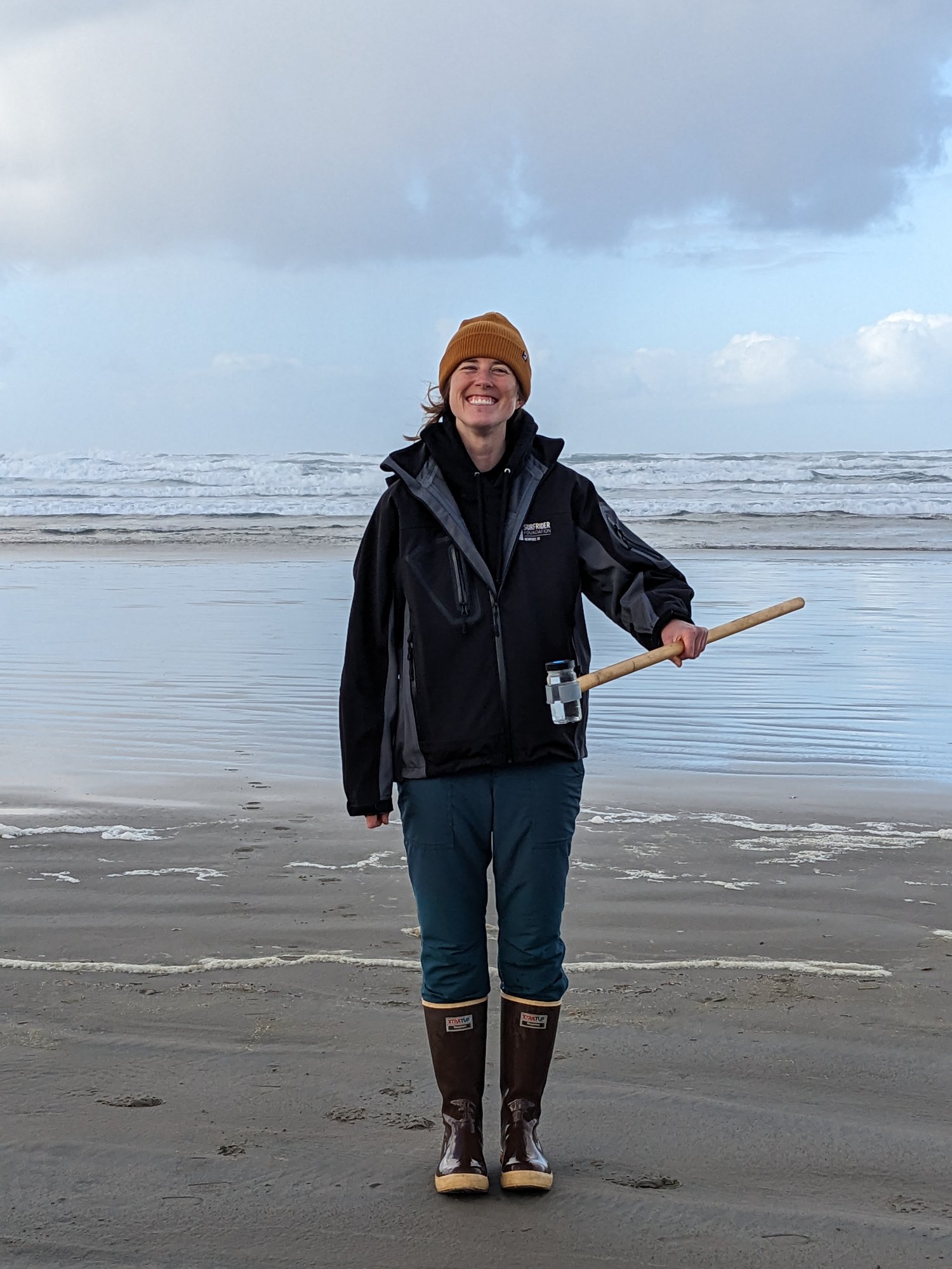 a smiling volunteer standing on Nye Beach.  She is holding a water sample in a jar at the end of a long wooden pole