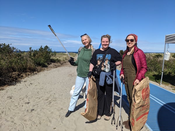three women pose with trash pickers and bags at a beach cleanup in Seaside