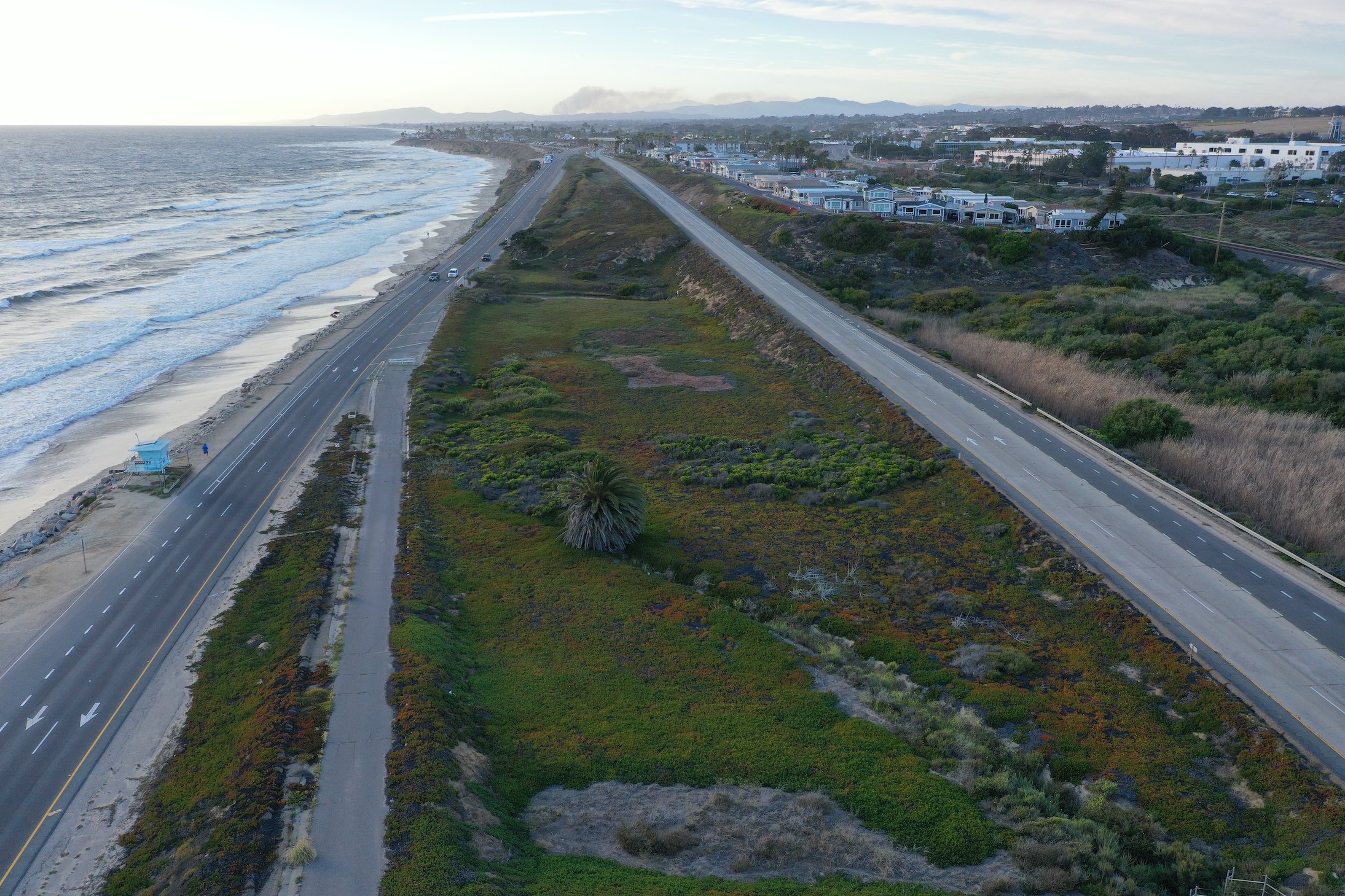 a drone image of the dip at Encinas Creek, showing the ocean encroaching on the southbound lane and a wide, undeveloped median between the south and northbound lanes.