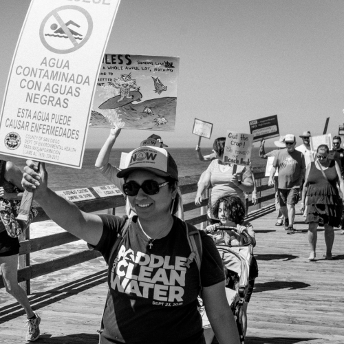 A black and white photo of protesters walking up a pier holding signs with messages about clean water. The main person in the image is a latina woman with light skin, sunglasses, a small crusafix necklace, a backpack, a hat that reads