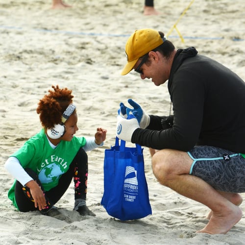 a child crouches down to pick up a peice of trash on a sandy beach. The child has dark skin, dark brownish reddish hair, and is wearing a bright green shirt with a cartoon image of the earth, and white noise reducing over the ear headphones. An adult crouches nearby holding open a blue trash collection back with the Surfrider wave logo. The adult is light skinned, wearing a yellow baseball hat and white and blue protective gloves.