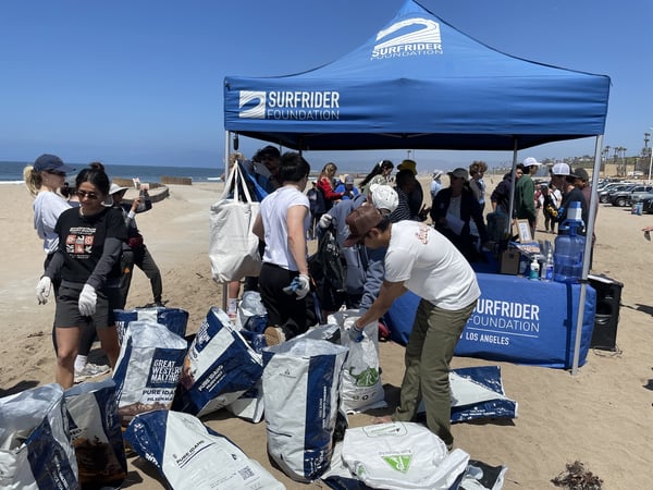 Surfrider Volunteers gather trash in big white bags around a branded Surfrider blue tent. 