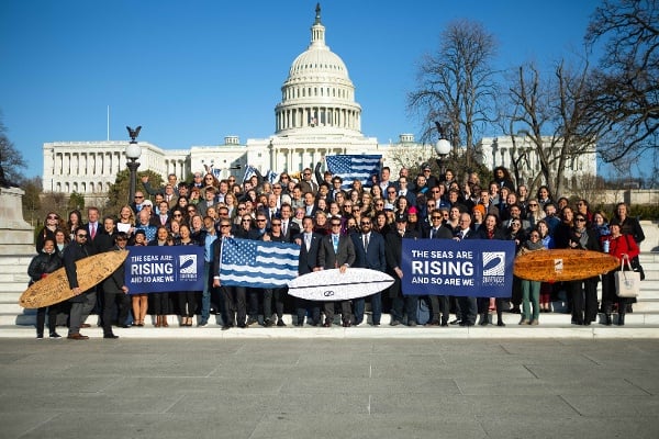 Dozens of volunteers pose in front of the US Capital building, holding up surfboards, the united oceans flag, and signs that say the seas are rising and so are we