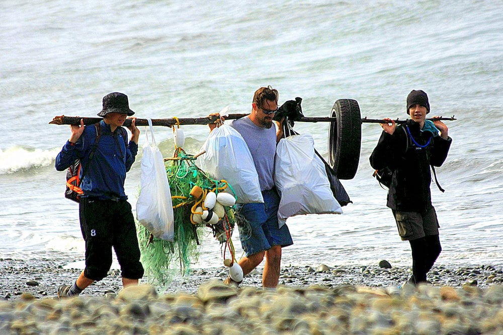 Three volunteers hold a long branch across their backs with trashbags, buoys, discarded rope, and a tire slung between them