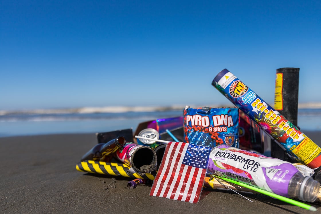 A pile of used fireworks and a small plastic American flag on the beach with the ocean in the background