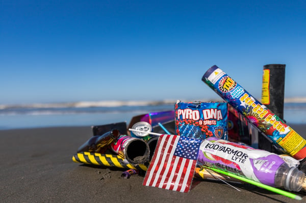 A pile of used fireworks and a small plastic American flag lying on the beach with the ocean in the background