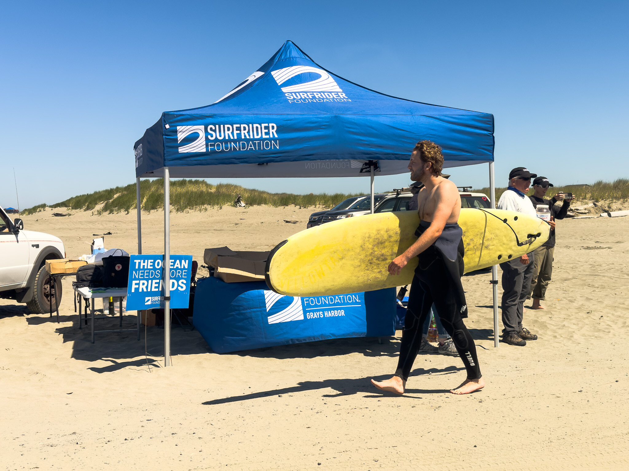 A volunteer walking past the Surfrider tent carrying a large yellow surfboard 