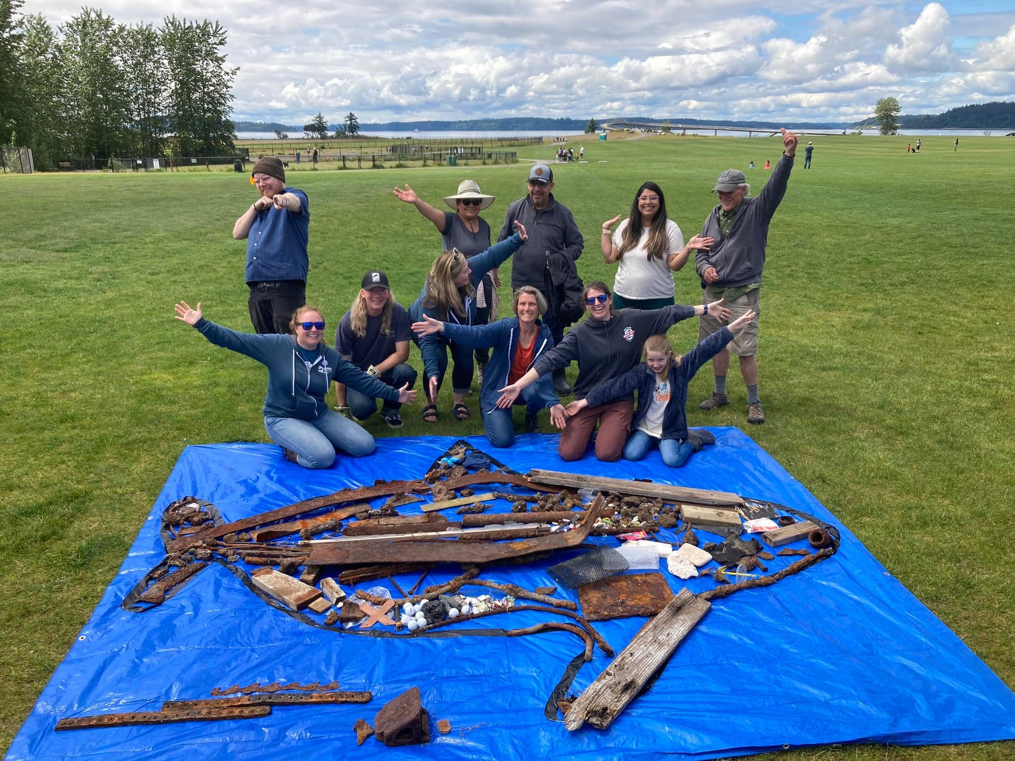 A dozen volunteers pose, arms out, by a large salmon figure assembled from debris on a blue tarp