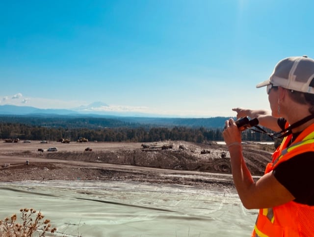 Wearing a high viz vest, Liz points to the active landfill beneath her on a hill, with Mt. Rainier in the distance 