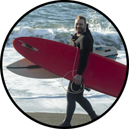 Pete wearing a wetsuit carrying a surfboard under each arm as he walks down the beach