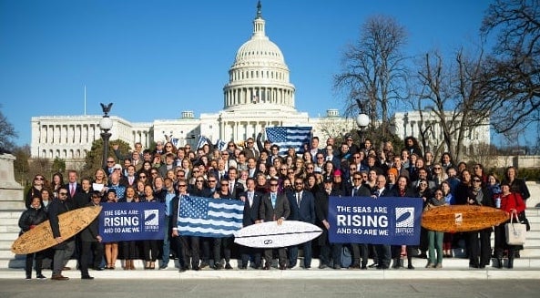 Dozens of Surfrider volunteers and staff pose in front of the US Capital building holding surfboards and signs