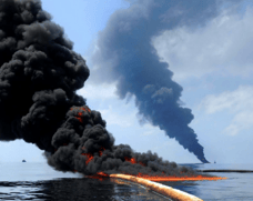 An oil spill on fire emitting black smoke against a blue sky
