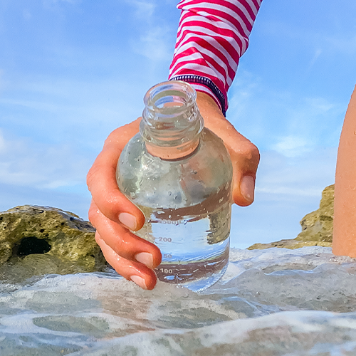 A close up from water level of a hand reaching down to pull a glass bottle up out of the surf. The background shows a blue sky, a big brown rock, and the edge of the beach cliffs.