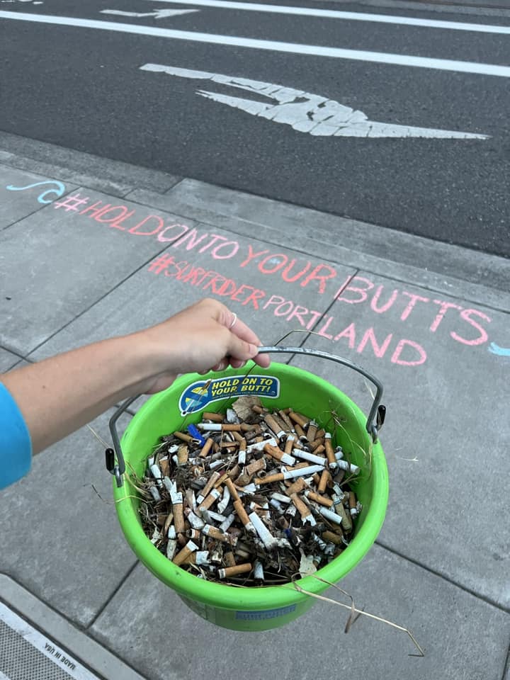 a volunteer holds a green bucket full of cigarette butts, with chalk writing in the background on the sidewalk reading "#holdontoyourbutts #surfriderportland