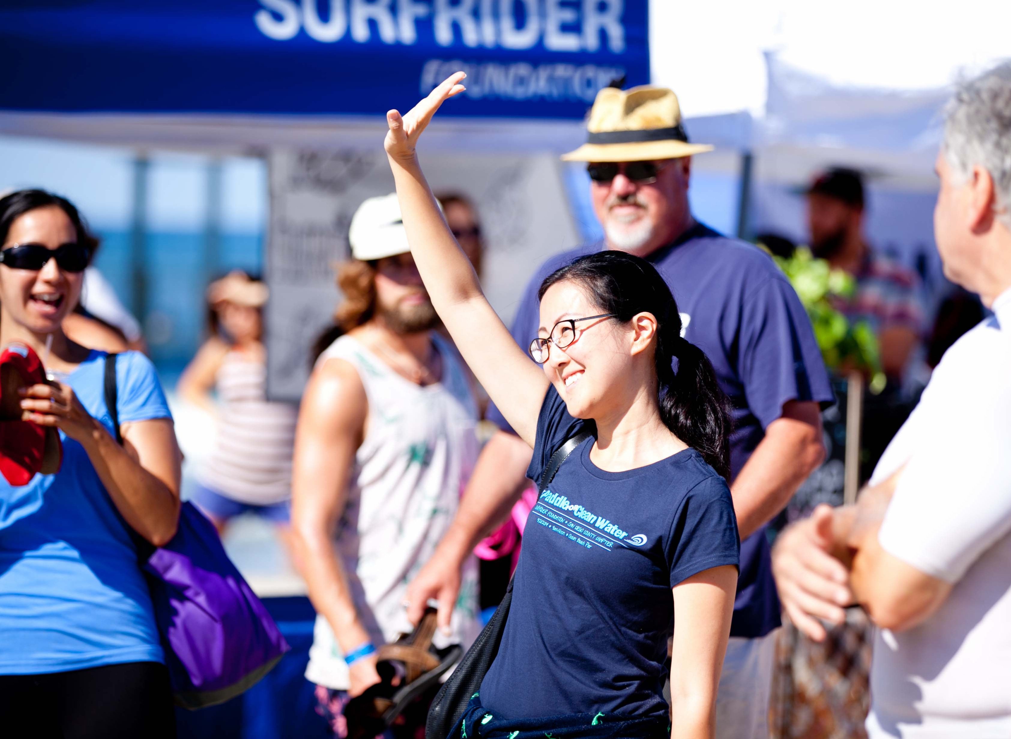 A woman raises her hand in the middle of a crowd of excited volunteers. The woman is wearing a dark blue paddle for clean water shirt with glasses and black hair
