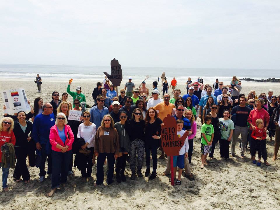 A large group of people gather at the beach with signs saying veto Port Ambrose