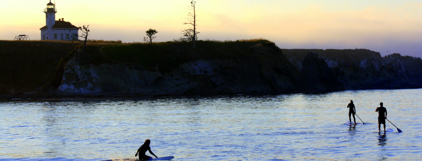 Three people are out on the water during sunset with a light house in the background. Two are stand up paddle boarding